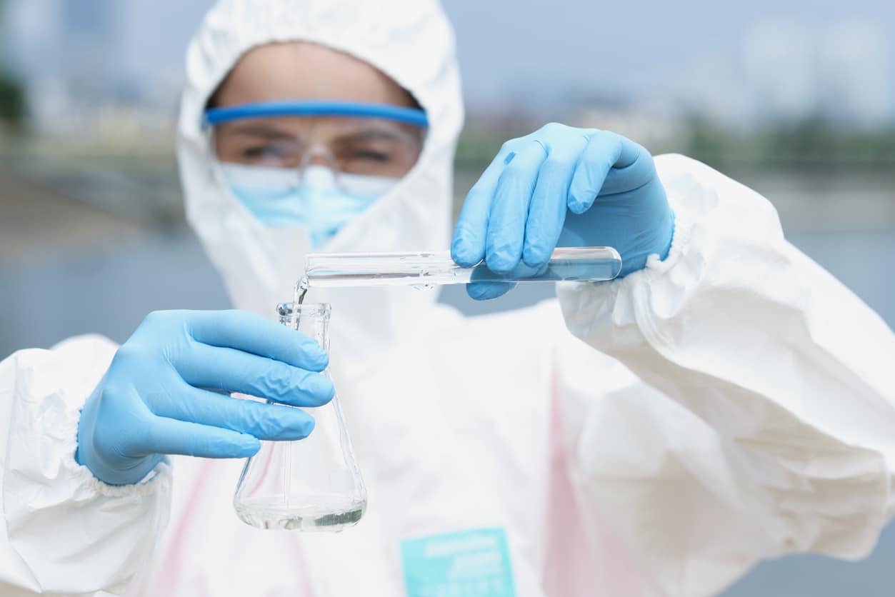 Researcher in protective suit and glasses with gloves pours water from test tube glass beaker. Water quality analysis concept